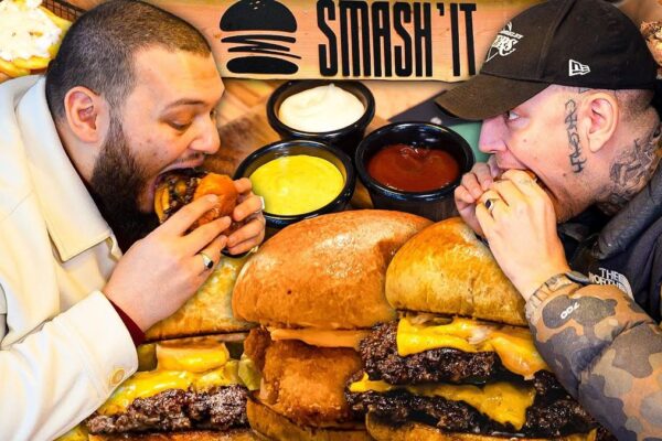 Alt-Text: "Video-Thumbnail von AbuGokus Video 'MONTE ZEIGT MIR DEN BESTEN BURGER IN BUXTEHUDE'. Das Bild zeigt AbuGoku und Monte vor einem Burger-Restaurant in Buxtehude, beide lächelnd und mit einem Burger in der Hand. Der Hintergrund zeigt das Restaurant-Schild und einige Passanten, die vorbeigehen.